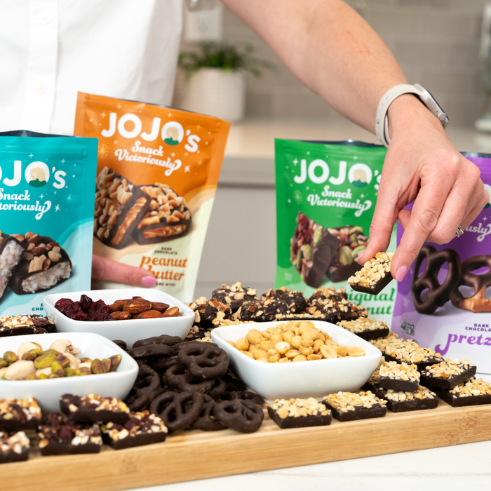 Best sellers dark chocolate bites on a charcuterie board. Woman's hand holding a peanut butter chocolate bite. 4 flavors, pistachio almond cranberry, peanut butter, coconut almond and chocolate covered pretzels.