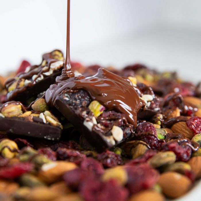 Dark chocolate pouring onto a stack of dark chocolate snacks with cranberries pistachios and almonds