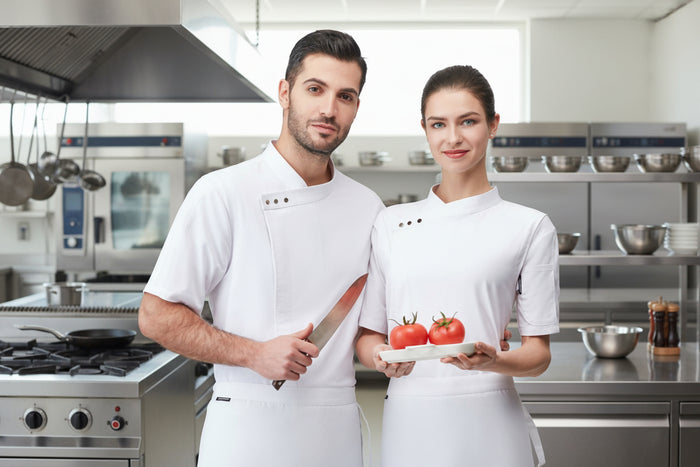 A male and a female chef wearing white overalls, with the male holding a knife and the female holding a tray with two tomatoes.