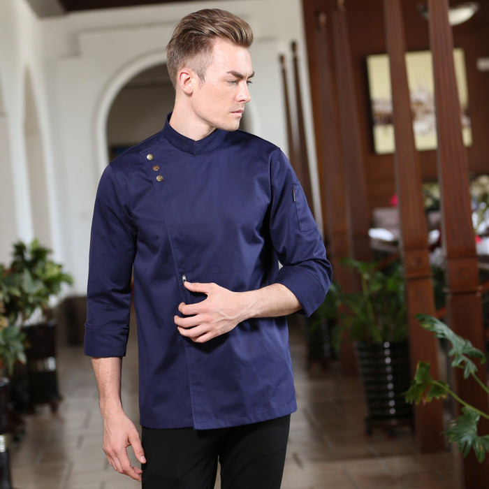 A male model wearing a long sleeve chef jacket in blue color with a mandarin collar, featuring front button closure and a left chest pocket.