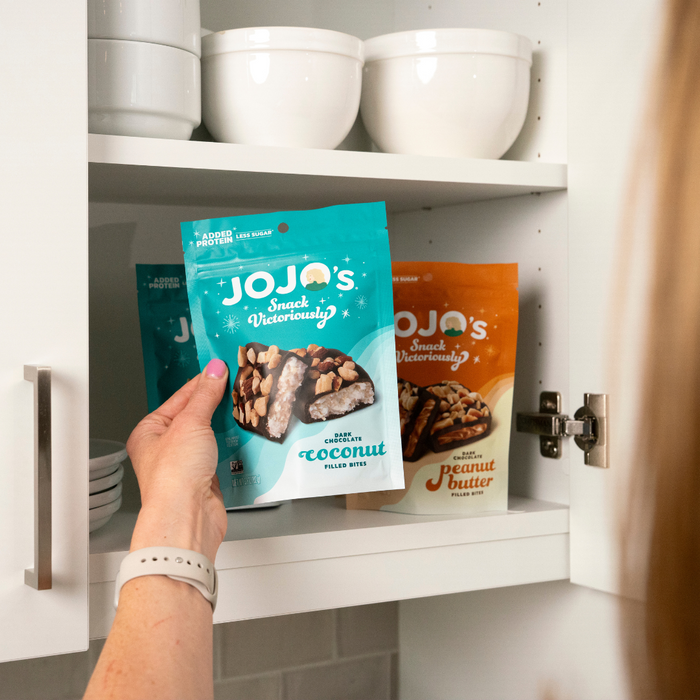 Hand holding a package of JOJO's snack in front of a kitchen cupboard with more packages.