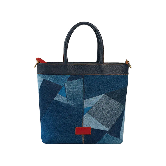 A structured tote bag featuring a blue geometric patchwork pattern with dark blue leather trim, double top handles, and small red accent tabs on the front.