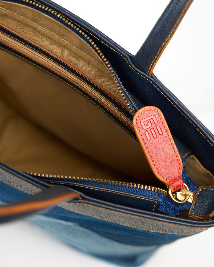 Close-up of a blue leather handbag interior showing a tan-lined compartment, gold zipper teeth, and a small orange-brown leather pull tab with a gold clasp.